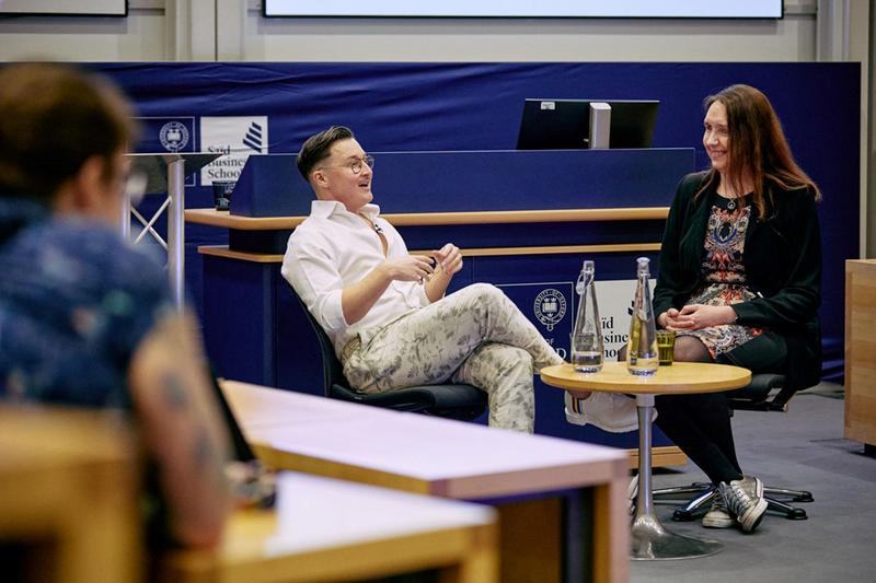 Sarah and Max are in conversation, smiling and laughing, sitting around a small table. They are both dressed smartly- Max in a white shirt and cream and green patterned trousers, and Sarah in a multicoloured dress with a black blazer. 